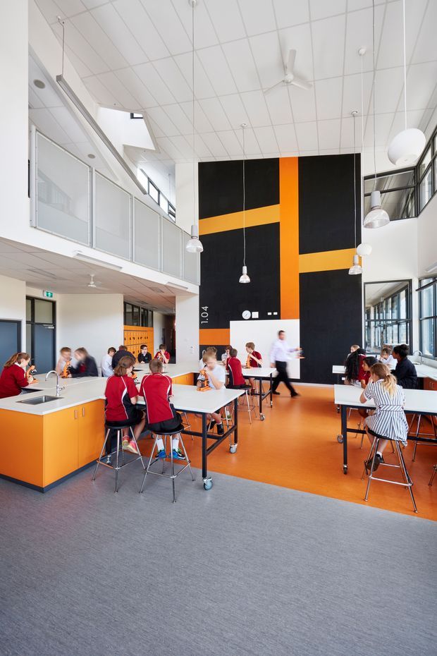 Bright, double-height school dining or common area with students seated at communal tables. The space features bold orange-and-black graphic wall panels, orange fixed cabinetry and flooring, grey carpet, extensive glazing, a mezzanine-level walkway, suspended pendant lights, and a gridded acoustic ceiling with ceiling fans.