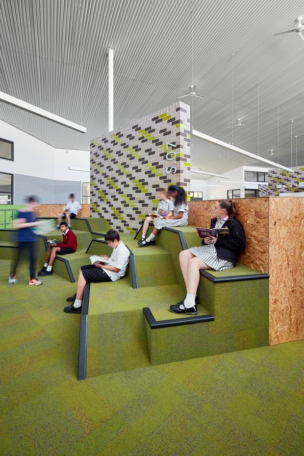 Bright, spacious school or educational interior with tiered green carpeted seating and integrated black-edged benches. Students are gathered in the informal learning area. The space features a high ribbed white ceiling with suspended linear lighting and ceiling fans, pale walls, timber-look OSB partition panels, and a prominent patterned brick feature wall with white, grey, black, and lime-green accents.