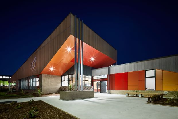 Night-time exterior view of a contemporary institutional or commercial building with a dramatic cantilevered entrance canopy. The facade combines dark vertical metal cladding, warm timber-toned panels, exposed brick, vivid red and orange soffit and wall panels, extensive glazing, and tall silver structural columns. A clearly marked reception entrance opens onto a broad paved forecourt with integrated planter beds and bench seating.