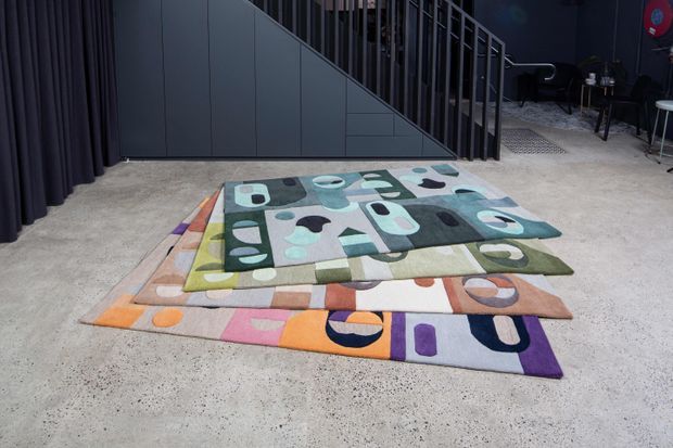 A dark, contemporary showroom or commercial interior with a polished concrete floor, charcoal wall panels, black curtains, and a metal stair railing. Several colourful, abstract patterned rugs are layered and displayed across the foreground floor. A small seating area is visible in the background.