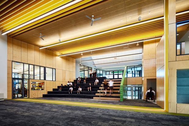 Large contemporary institutional interior featuring a stepped black seating platform or informal auditorium in a tall open lobby. The space has extensive warm timber wall and ceiling panelling, linear timber ceiling battens with integrated lighting, black-framed glazing, polished concrete or dark carpeted flooring, and a prominent green stair or ramp element. People are seated and walking through the space.