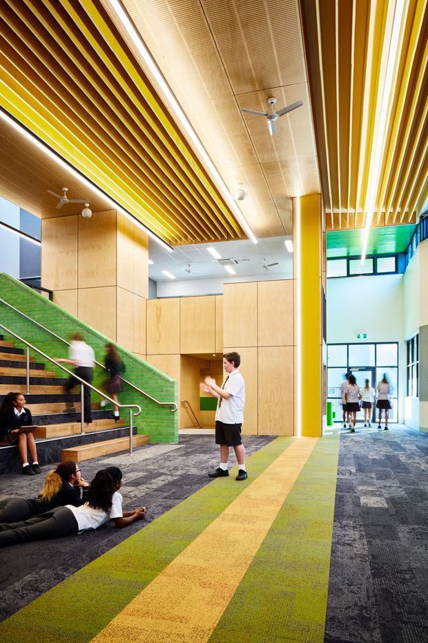 A brightly lit multi-level institutional interior, likely a school or educational facility, viewed from a spacious circulation area. The space features broad carpeted flooring in grey, green and yellow bands, timber-panelled walls, a green tiled stair riser and balustrade zone, vivid yellow structural columns, extensive timber slatted ceiling features with integrated linear lighting, ceiling fans, and large glazed openings. Students occupy the stairs and floor, with a corridor extending toward the rear.