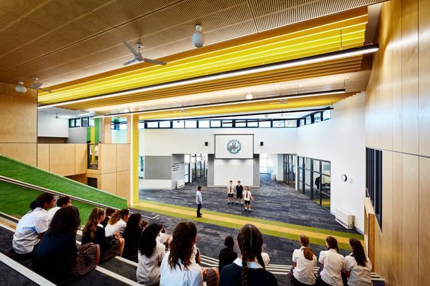 A contemporary multi-level educational or institutional interior viewed from tiered auditorium-style seating. Students sit in the foreground facing an open central atrium with a broad green sloped ramp, white walls, glazed corridors, timber wall and ceiling panels, and long linear yellow ceiling light or acoustic elements. The space has a large, bright, communal and highly designed character.