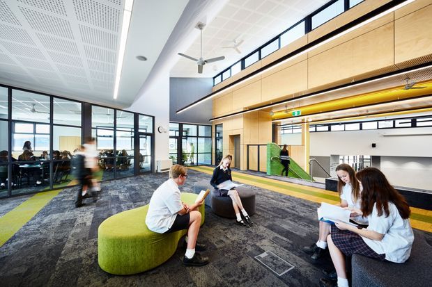 A bright, contemporary multi-level commercial interior, likely an educational or institutional common area. The open lounge has students seated on rounded upholstered ottomans and benches, a large patterned carpet, extensive internal glass partitions, and a tall timber-lined atrium with a mezzanine or upper-level walkway. Linear illuminated ceiling features, exposed services, ceiling fans, and dark-framed glazing contribute to a modern collaborative environment.