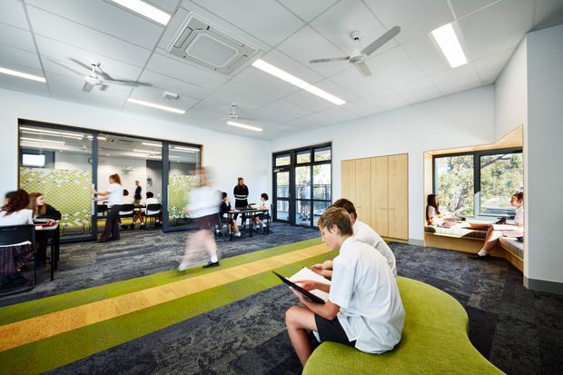 A bright contemporary communal interior, likely a school or educational lounge, with students gathered around tables and seating. The space has a patterned dark carpet, a large curved lime-green and natural-wood-toned built-in bench, extensive black-framed glazing, timber wall panels, white acoustic ceiling tiles, linear LED lighting, ceiling fans, and an air-conditioning cassette. A kitchenette or breakout area is visible through the glazed partition.