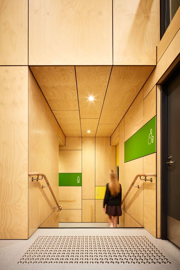 A compact, highly finished interior corridor or entrance to a bathroom facility, lined floor-to-ceiling with warm plywood panels and exposed dark joints. The ceiling continues the timber panelling and contains recessed spotlights. Stainless-steel handrails run along both side walls, while bright green accessibility and restroom signage provides colour contrast. A textured tactile entrance mat occupies the foreground, and a dark door is visible at right.
