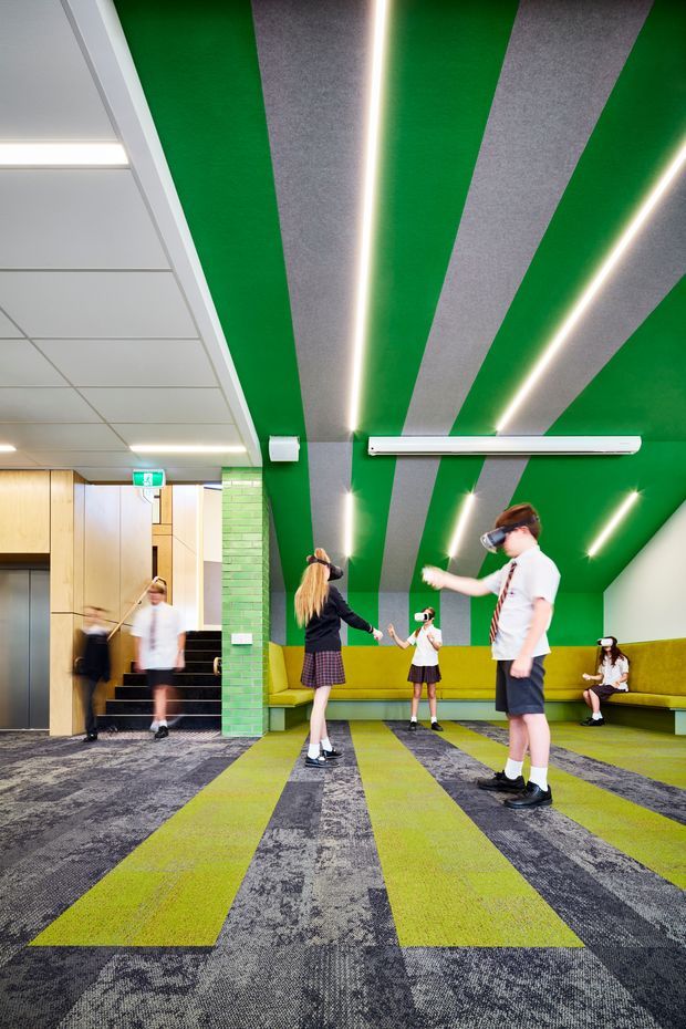 Vibrant contemporary commercial interior, likely a school or youth activity space, with a green-and-grey striped ceiling and matching patterned carpet. Linear LED lighting follows the ceiling bands, while lime-green upholstered bench seating lines the walls. A tiled green column, stair and lift lobby, emergency exit signage, and children wearing virtual-reality headsets are visible.