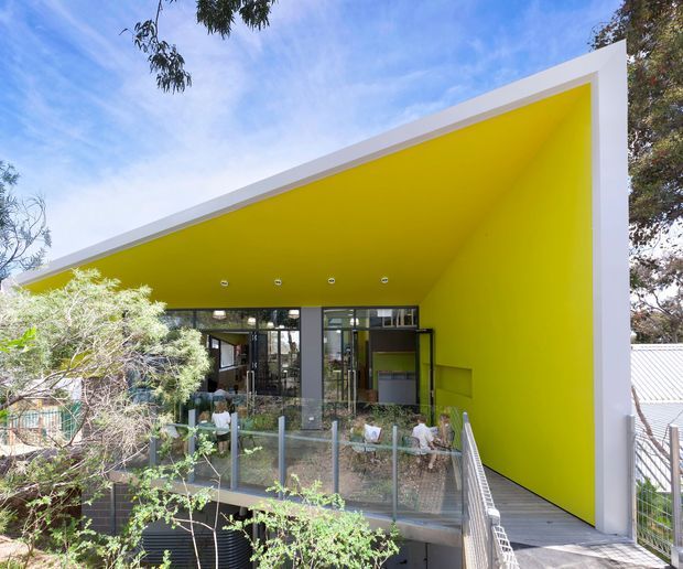 Exterior view of a contemporary single-storey building with a dramatic angular mono-pitched roof and a vivid yellow recessed soffit and side wall. Extensive glazing opens onto a raised deck or terrace with metal balustrades, surrounded by established trees and planting.