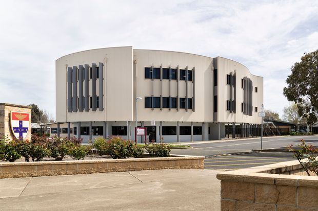 Exterior view of a contemporary multi-storey institutional or commercial building with a sculptural curved facade, pale metal cladding, dark vertical sun-shading fins, and rows of dark-framed windows. The building is set within a landscaped campus with paved pedestrian areas, planted beds, and a roadway.