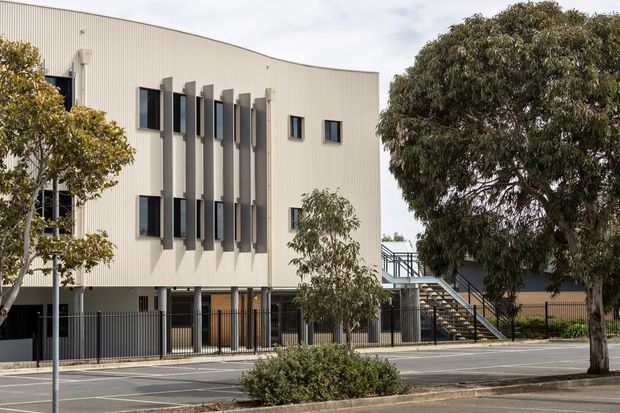 Street-facing view of a contemporary multi-storey building with a pale ribbed metal-clad facade, vertically projecting window surrounds, dark-framed windows, ground-level glazing and an external stairway. Mature street trees and marked on-street parking occupy the foreground.
