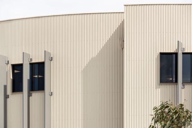 Exterior close-up of a contemporary building facade clad in pale cream vertically ribbed profiled panels. Dark-framed windows are paired with projecting vertical fins or screening elements, creating strong geometric shadows in bright sunlight. A small portion of landscaping is visible at the lower edge.