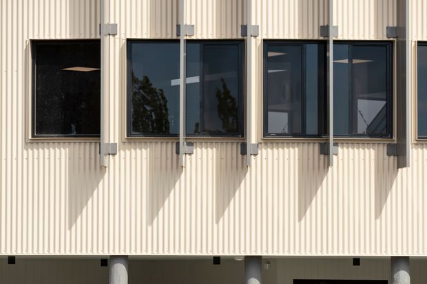 Close-up exterior view of a contemporary building facade clad in pale vertically ribbed metal or corrugated panels. A row of dark-framed rectangular windows is fitted with prominent external vertical support or sun-shading elements, casting strong geometric shadows across the facade. Partial structural columns and openings are visible along the lower edge.