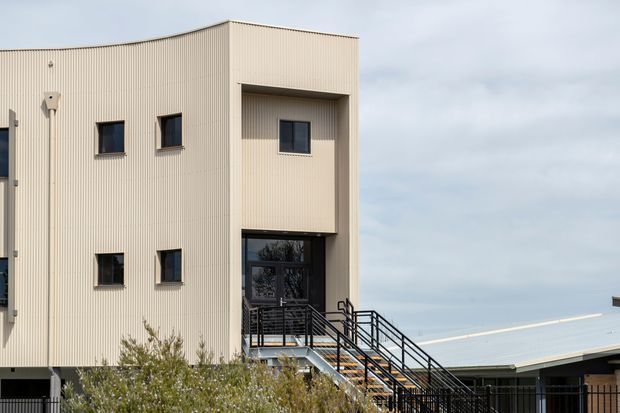 Exterior view of a contemporary multi-storey building with pale ribbed metal cladding, dark-framed windows, recessed upper-level glazing, and an exposed black metal staircase and balustrade. A low adjoining structure with a light metal roof is visible to the right, with landscaping in the foreground under an overcast sky.