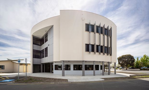 Exterior view of a modern multi-storey commercial building with a distinctive curved, pale facade. The building features vertical fin-like sun-shading elements, dark horizontal window bands, an elevated ground level supported by slender columns, and a paved street frontage with accessible parking nearby.