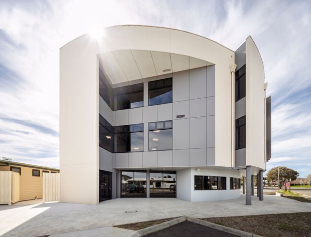 Exterior view of a contemporary multi-storey commercial building with a curved white facade, large dark-framed glazing, grey panel cladding, a recessed upper-level void, and slender structural columns. The building fronts a paved forecourt and appears to sit in a suburban or institutional setting.