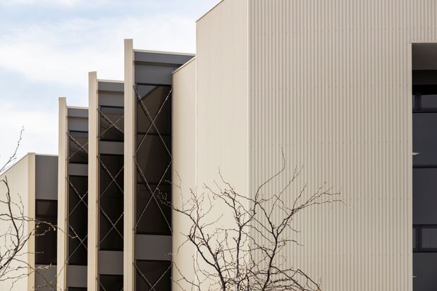 Exterior architectural photograph of a contemporary multi-storey building. The facade combines tall pale ribbed metal cladding with recessed dark glazed or screened bays, exposed vertical structural elements, and visible cross-bracing. Leafless branches partially obscure the lower foreground.