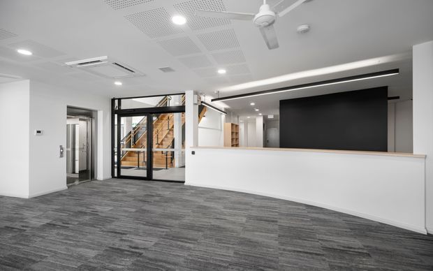 Bright contemporary commercial reception area with a long white reception desk, dark feature wall, grey carpet tile flooring, black-framed glazed entry doors, and a visible timber staircase beyond. The space has a suspended acoustic ceiling with recessed lighting, linear feature lighting, and a ceiling fan.