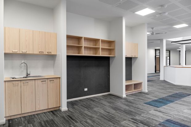 A bright contemporary commercial office interior with a compact kitchenette, light timber cabinetry, open built-in shelving, dark feature wall panels, and grey patterned carpet tile flooring. White structural columns, a suspended acoustic ceiling with recessed lighting, and a reception or service counter are visible in the background.