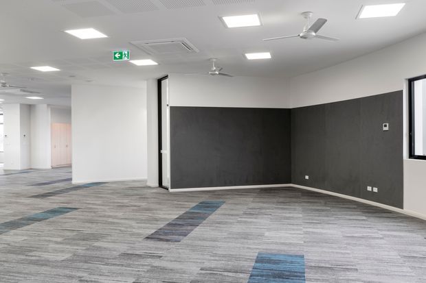 An empty, modern commercial office interior with grey patterned carpet tiles and occasional blue accent strips, white walls, a dark charcoal feature wall, recessed rectangular ceiling lights, exposed air-conditioning vents, ceiling fans, and a window. An open circulation area and exit signage are visible to the left.