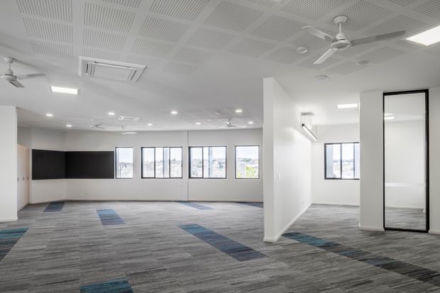 Bright, open-plan commercial interior with a carpeted floor, white walls, exposed acoustic ceiling tiles, multiple ceiling fans, recessed lighting, air-conditioning, and large windows. A black feature wall and partial-height partitions define flexible workspace or meeting areas.