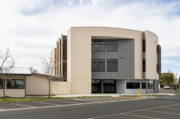 Exterior photograph of a contemporary multi-storey institutional or commercial building beside a marked parking area. The building has a pale ribbed metal-clad curved facade, darker rectangular panel sections with horizontal strip windows, a recessed glazed ground-level entry, and a projecting vertical side element. A lower adjoining weatherboard-clad structure is visible at left.