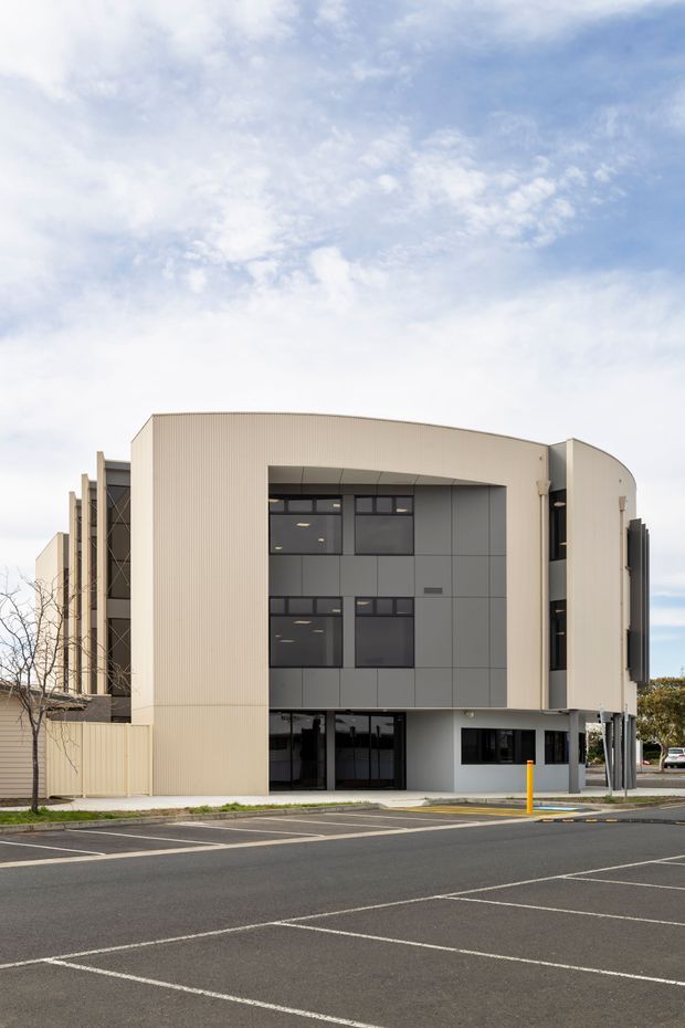 Exterior view of a contemporary multi-storey commercial building with a curved cream ribbed-metal facade, dark grey panel cladding, extensive dark-framed glazing, and a recessed ground-floor entry. The building fronts a marked asphalt parking area and includes projecting side volumes and vertical architectural fins. Bright overcast sky and sparse landscaping surround the structure.