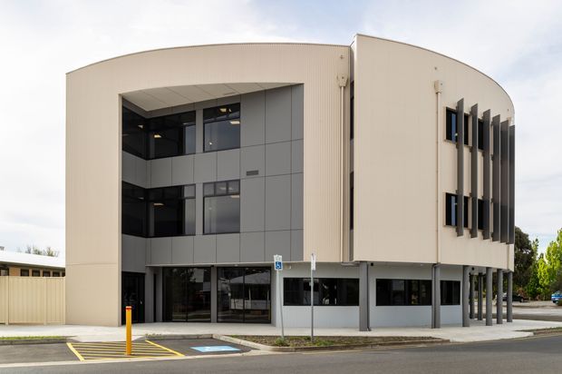 Exterior view of a contemporary multi-storey commercial building with a rounded, ribbed light-coloured facade, dark grey panel cladding, extensive glazing, and a covered ground-level entry supported by slender columns. Accessible parking and a paved street frontage are visible.