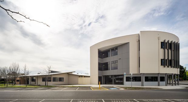 Exterior street-level view of a contemporary multi-storey institutional or commercial building with a distinctive curved corner facade, vertical fins, dark-framed windows, and grey panel cladding. A lower single-storey wing extends to the left, with accessible parking and a paved road in the foreground.