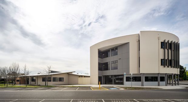 Exterior streetscape view of a contemporary multi-storey institutional or commercial building with a rounded cream-coloured facade, dark vertical sun-shading fins, extensive glazing, and a covered ground-level entry. A lower flat-roofed wing extends to the left, with marked parking and accessible parking spaces in the foreground.