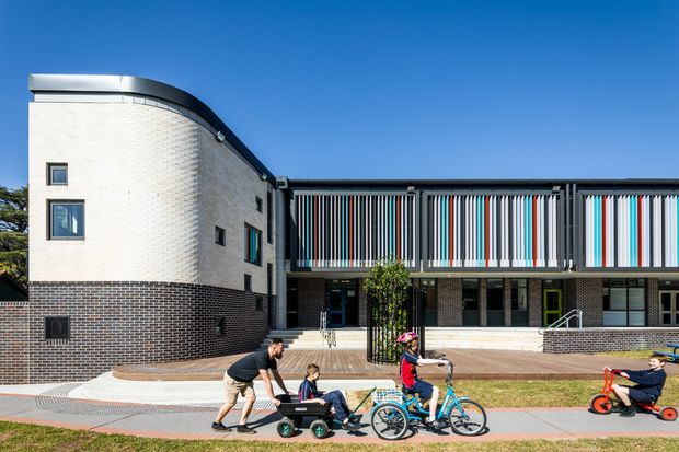Exterior view of a contemporary multi-storey building with a rounded white-brick corner wing and a long dark-brick facade. The upper level features striking vertical fins in alternating grey, white, blue and red tones, while a broad paved forecourt and accessible entry stairs connect to the pedestrian path. People and bicycles are present in the foreground, with clear blue sky and landscaping around the building.