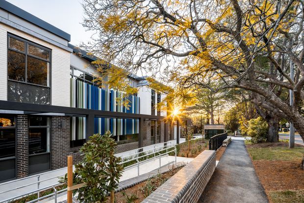 Street-facing view of a contemporary multi-unit building with dark brickwork, light rendered walls, large black-framed windows, and a distinctive vertical blue and grey screen along the upper facade. A ramp with metal handrails leads beside a low brick boundary wall to a paved pedestrian path, with mature street trees lining the verge.