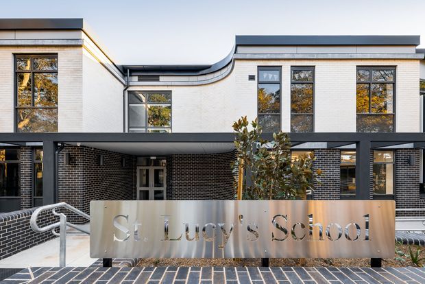 Front exterior of St Lucy's School, showing a contemporary low-rise institutional building with pale brick walls, dark brick lower sections, large multi-pane windows, dark metal rooflines and canopy framing, curved roof detailing, and a metal school sign in the foreground.