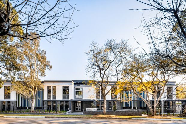 Street-facing exterior view of a contemporary multi-unit residential building, with a long low-rise facade, white rendered panels, dark brickwork, extensive glazing, and mature deciduous trees along the pavement. The building has a mostly flat roofline and a strong horizontal composition.