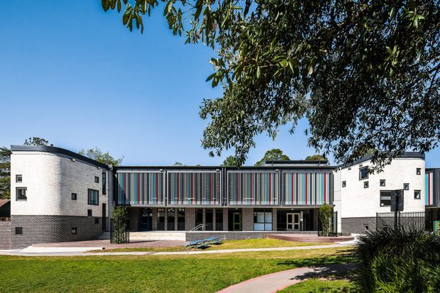 Front exterior view of a contemporary multi-storey building with curved white masonry end wings, dark brick lower sections, and a prominent central facade featuring vertical multicoloured fins or screens. The building is set behind a landscaped lawn with curved paved paths and mature trees under a clear blue sky.