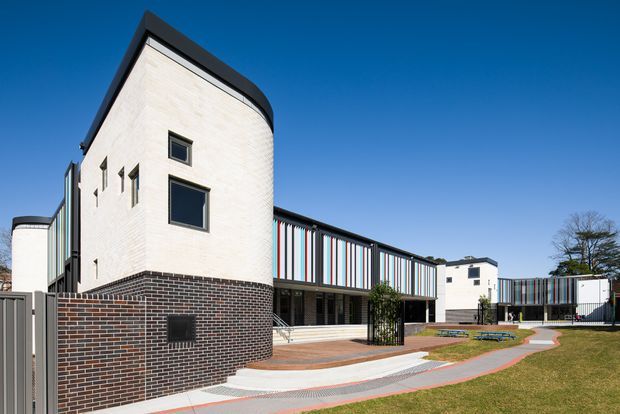 Exterior view of a contemporary multi-building complex with curved cream masonry walls, dark brick lower sections, black roof edges, and colourful vertical facade fins. Paved pedestrian paths and landscaped lawns surround the building under a clear blue sky.