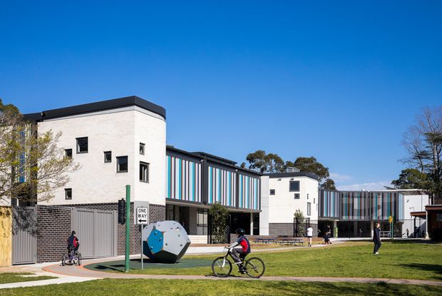 Exterior view of a contemporary multi-building institutional or educational complex under a clear blue sky. The development combines pale masonry volumes with dark trim, extensive vertically striped multicolour cladding, small square windows, landscaped lawn, paved walkways, bicycle activity, and a sculptural outdoor play or recreation element.