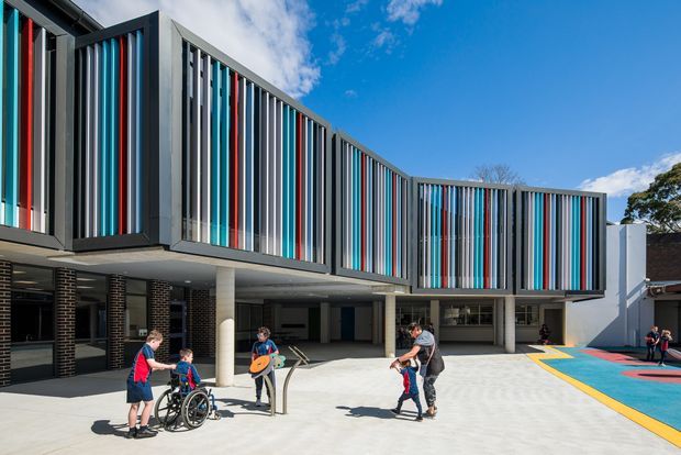 Exterior view of a contemporary multi-storey educational or community building with a dramatically curved facade clad in vertical fins. The fins alternate dark grey, white, turquoise, and red accents, creating a vivid striped elevation. A covered ground-level walkway with concrete columns opens onto a paved courtyard, with a colourful sports or play surface at the side. People are present for scale.
