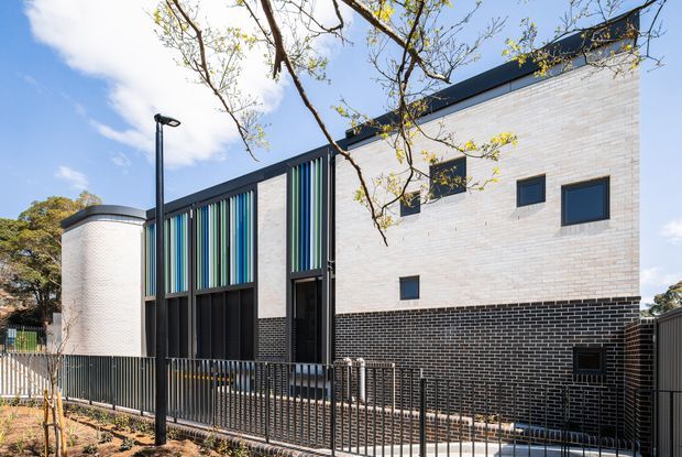 Exterior view of a contemporary multi-level building with a predominantly white brick facade, dark brick base, black metal roof edging, and vertical multicoloured screening elements around the entrance. The building has narrow rectangular windows, a curved corner wall, black metal fencing, and a landscaped street frontage.