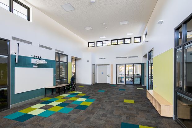 Bright, spacious institutional interior lobby or shared circulation area with a high ceiling, clerestory glazing, extensive internal windows, colourful geometric carpet tiles, white walls, teal wall panelling, and a lime-green built-in feature wall with bench seating. The space includes accessible circulation, glazed doors, and educational or healthcare-style functional fittings.
