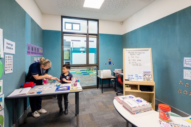 A bright, compact educational support or classroom space with teal acoustic wall panels, white upper walls, carpeted flooring, and a suspended acoustic ceiling. A teacher works with a child at a rectangular activity table, while educational posters, visual learning materials, a whiteboard on an easel, books, and storage furniture are arranged around the room. Large windows provide views into an adjacent school area.
