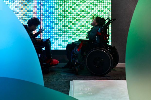 A dimly lit immersive interior or exhibition space with two people using wheelchairs, framed by large curved blue and green sculptural forms. A vibrant pixelated LED wall glows in blue, turquoise, green, and white behind them, while a pale illuminated or reflective rectangular surface occupies the foreground. The scene has a contemporary, experiential, technology-focused atmosphere with clear accessibility relevance.