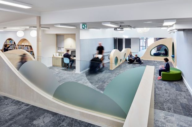 A bright contemporary commercial interior, likely a collaborative office or learning space, with open-plan work and lounge zones. The room features sculptural curved pale-wood and frosted-glass partitions, built-in pod-like seating, white ceilings with recessed linear lighting, patterned grey carpet, and colourful informal furniture. Several people are present, with desks, shelving, and a kitchenette or work area visible in the background.