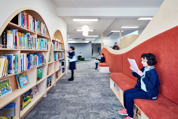 Bright contemporary school library or reading area with extensive built-in arched timber bookshelves on the left and curved red upholstered bench seating with integrated book storage on the right. Students are reading and moving through a spacious carpeted area beneath exposed ceiling beams and modern rectangular and globe lighting.