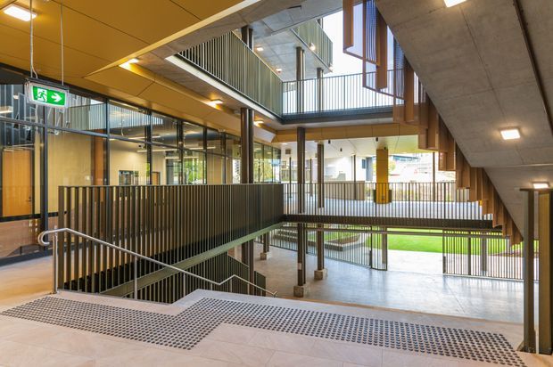 Interior view of a contemporary multi-level commercial or institutional building, showing an open circulation atrium with stairs, metal balustrades, exposed concrete, extensive glazing, timber-toned wall panels, and multiple walkways overlooking a landscaped courtyard. Emergency exit signage and strong natural daylight are visible.