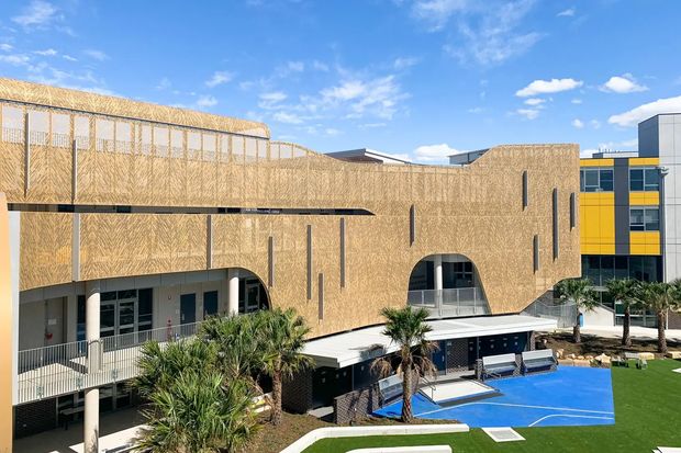 Exterior view of a contemporary multi-storey educational or institutional complex surrounding a landscaped courtyard. The facade is predominantly clad in warm tan patterned panels with expressive arched openings, narrow vertical slit windows, covered walkways, balconies, and a flat roofline. A bright blue outdoor sports court, synthetic lawn, palm trees, and adjacent black-and-yellow facade elements are visible under a clear sky.