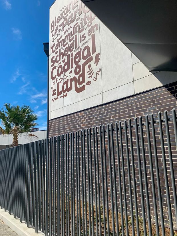 Exterior view of a contemporary educational building, featuring a dark brick wall, tall vertical metal security fencing, pale modular wall cladding, and a large typographic mural reading “Learning ... Creativity ... Coding ... Language.” A palm tree and blue sky are visible beside the building.