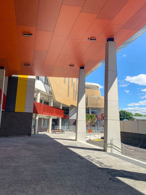 A bright outdoor architectural view beneath a large orange-toned soffit canopy supported by tall rectangular concrete columns. The paved forecourt includes stairs, metal handrails, benches, and a colourful building facade with black brickwork, yellow and red panels, curved beige cladding, and visible balconies. Blue sky and greenery appear beyond the structure.