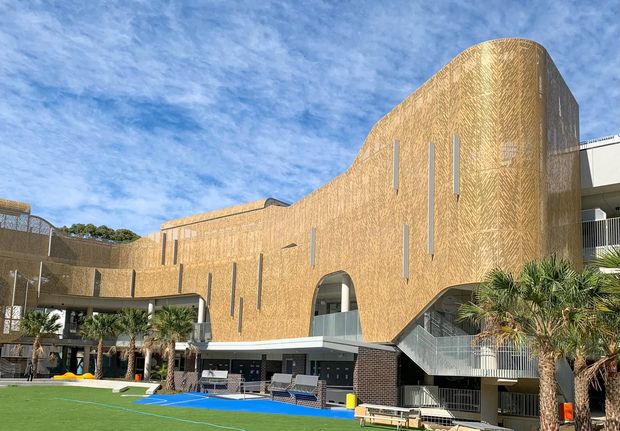 Exterior view of a large contemporary curved building with a distinctive golden-brown textured facade, narrow vertical slit windows, arched openings, external stairs, balconies, and a landscaped recreational area with palms and artificial turf in the foreground. The building appears institutional or commercial, with a bright blue pool or court-like surface at its base.