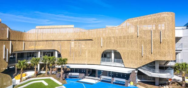 Exterior architectural view of a contemporary multi-storey building with a sweeping curved facade clad in a warm tan patterned material, narrow vertical windows, projecting balconies, and a lower-level pool courtyard with lounge areas and landscaping. Bright sunny conditions and a clear blue sky emphasize the building's sculptural form.