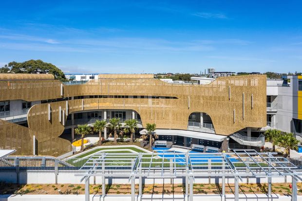 Aerial architectural view of a contemporary multi-storey building with extensive curved forms and a distinctive woven or perforated tan facade screen. The complex includes terraces, landscaped areas, a blue swimming pool, and a white pergola structure in the foreground, set within an urban or suburban environment under a clear blue sky.